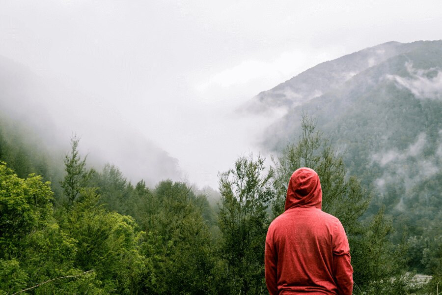 a man in a red sweatshirt stands in the rain in the mountains