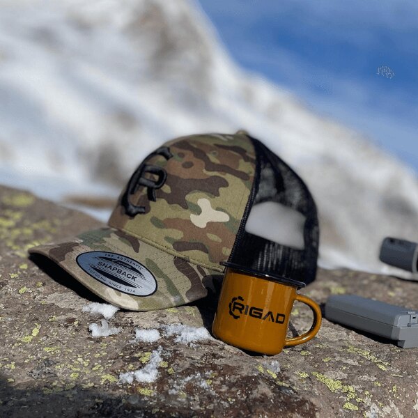 Rigad<sup>&reg;</sup> Retro Trucker FlexFit<sup>&reg;</sup> cap with the Rigad Logo tin mug in the mountains. Source: Rigad