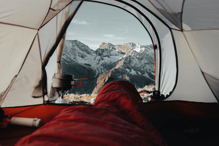 A view from a red sleeping bag of snow covered mountains. Source: https://www.pexels.com/cs-cz/foto/krajina-hory-cestovani-dobrodruzstvi-9166687/